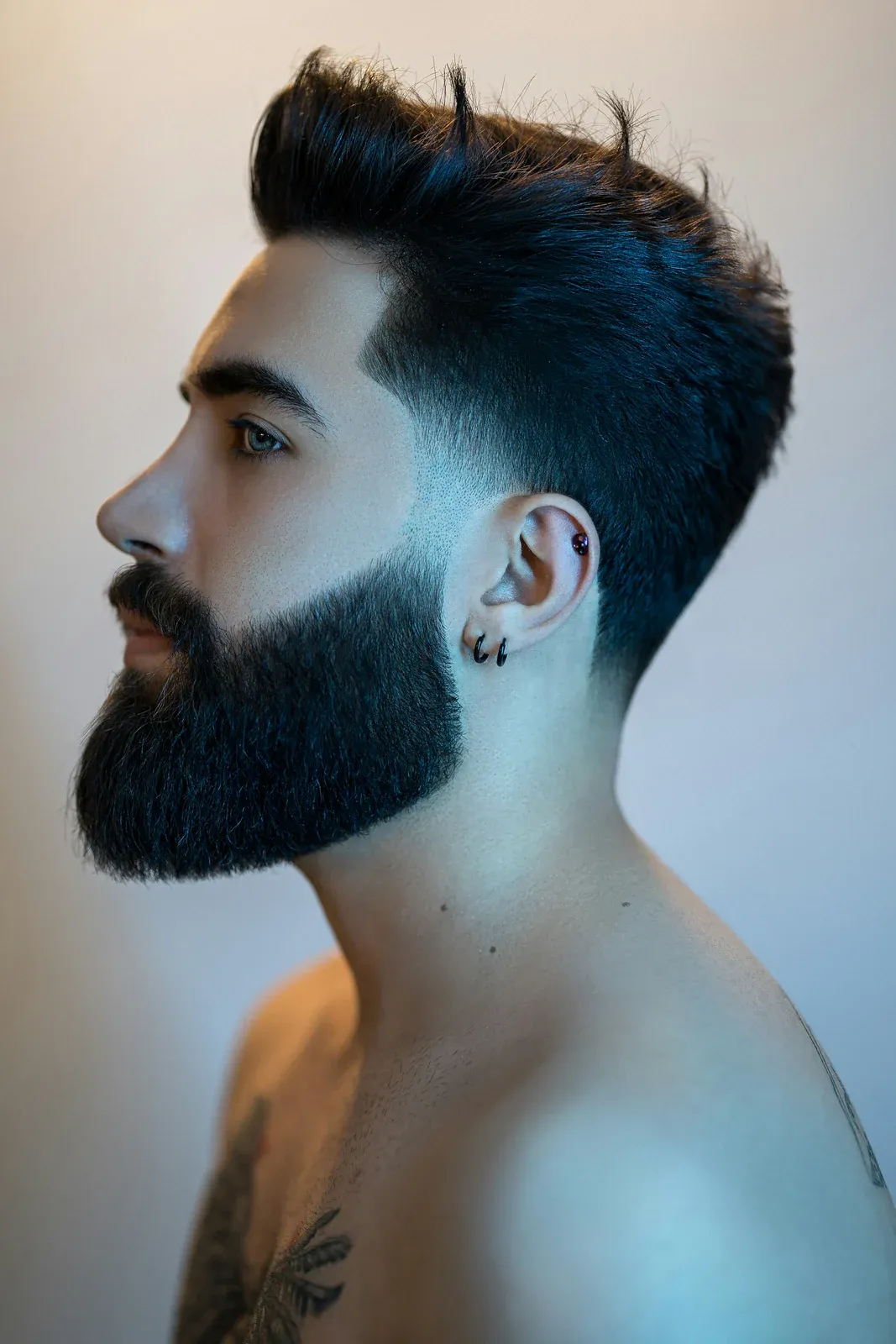 Homem de perfil com barba cheia escura que afina na costeleta e emenda num degradê no cabelo