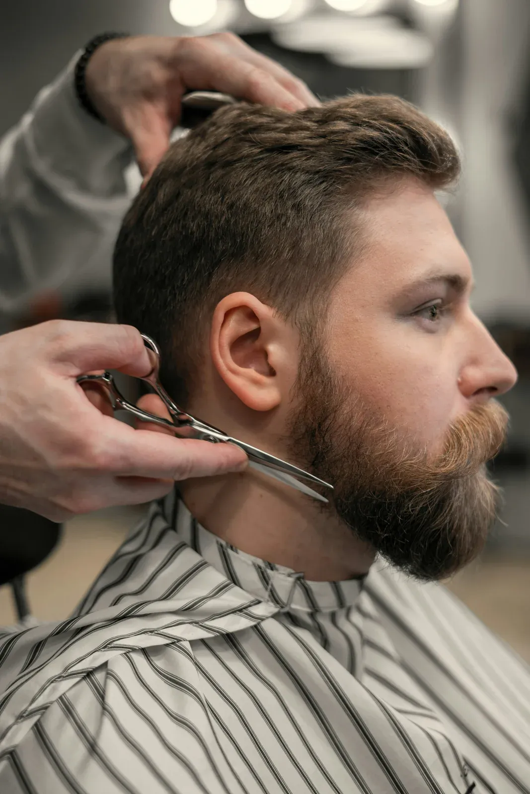 Barbeiro aparando com tesoura a lateral da barba ruiva de um homem de perfil, com capa listrada