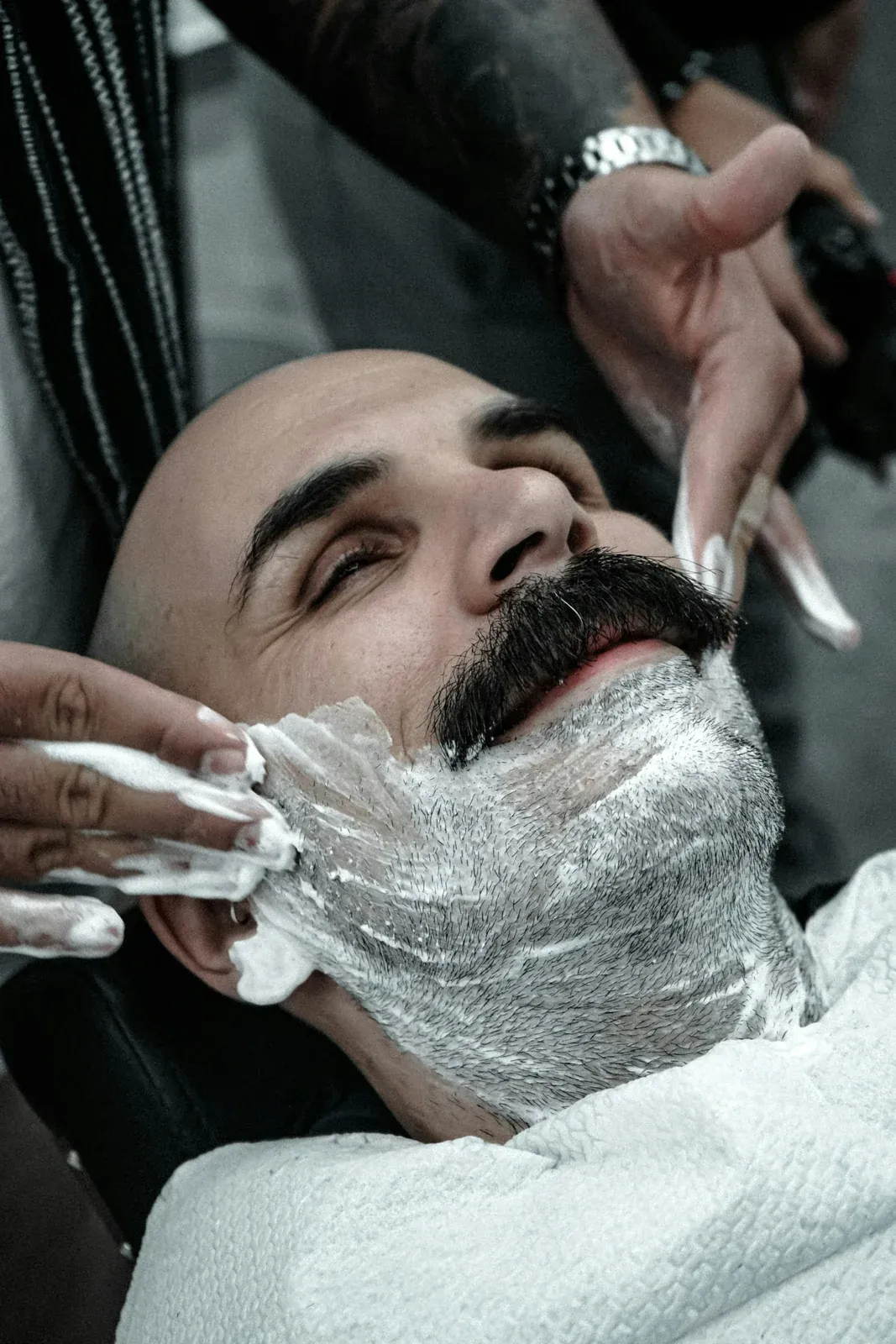 Homem de bigode reclinado na cadeira enquanto o barbeiro espalha espuma de barbear pelo queixo e pelo pescoço