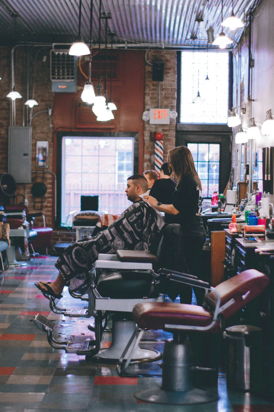 Interior de barbearia com cadeiras antigas e piso xadrez, onde uma barbeira atende um cliente coberto pela capa