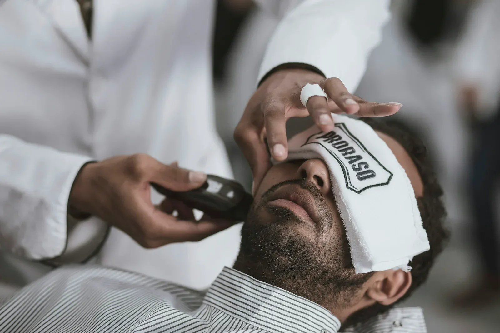 Homem reclinado na cadeira com uma toalha branca sobre os olhos enquanto o barbeiro passa a máquina na barba