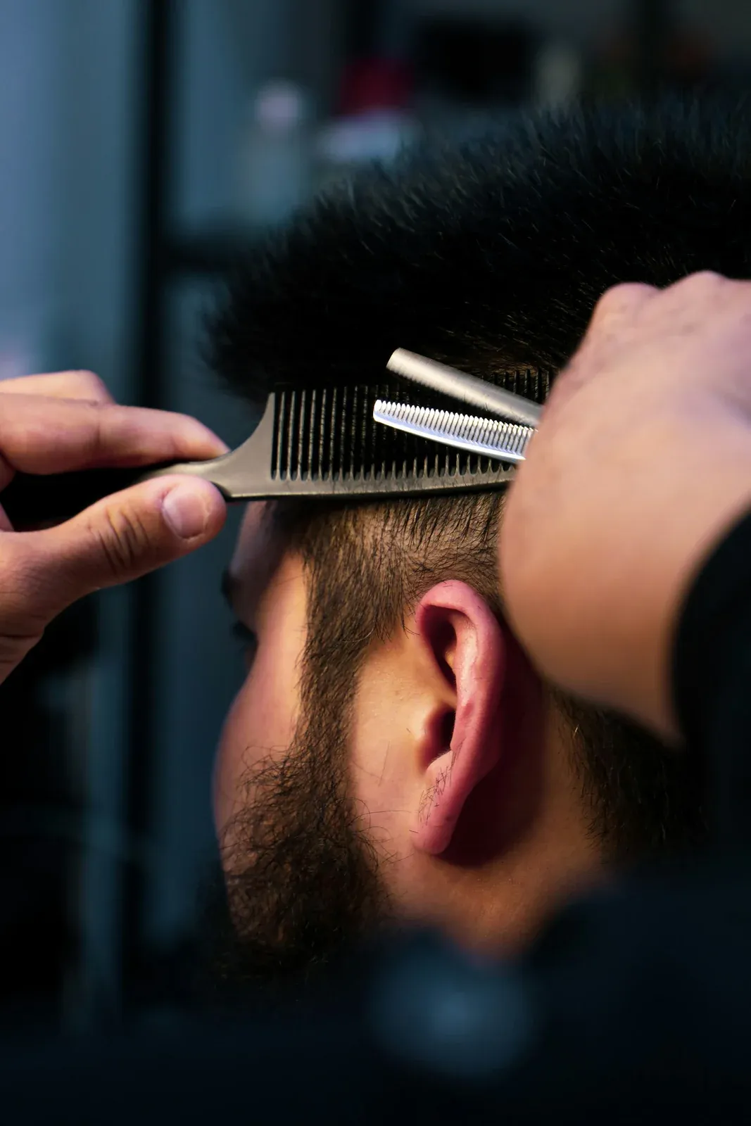 Barbeiro levantando o cabelo do topo com o pente e aparando com a tesoura de desbaste, acima da orelha do cliente